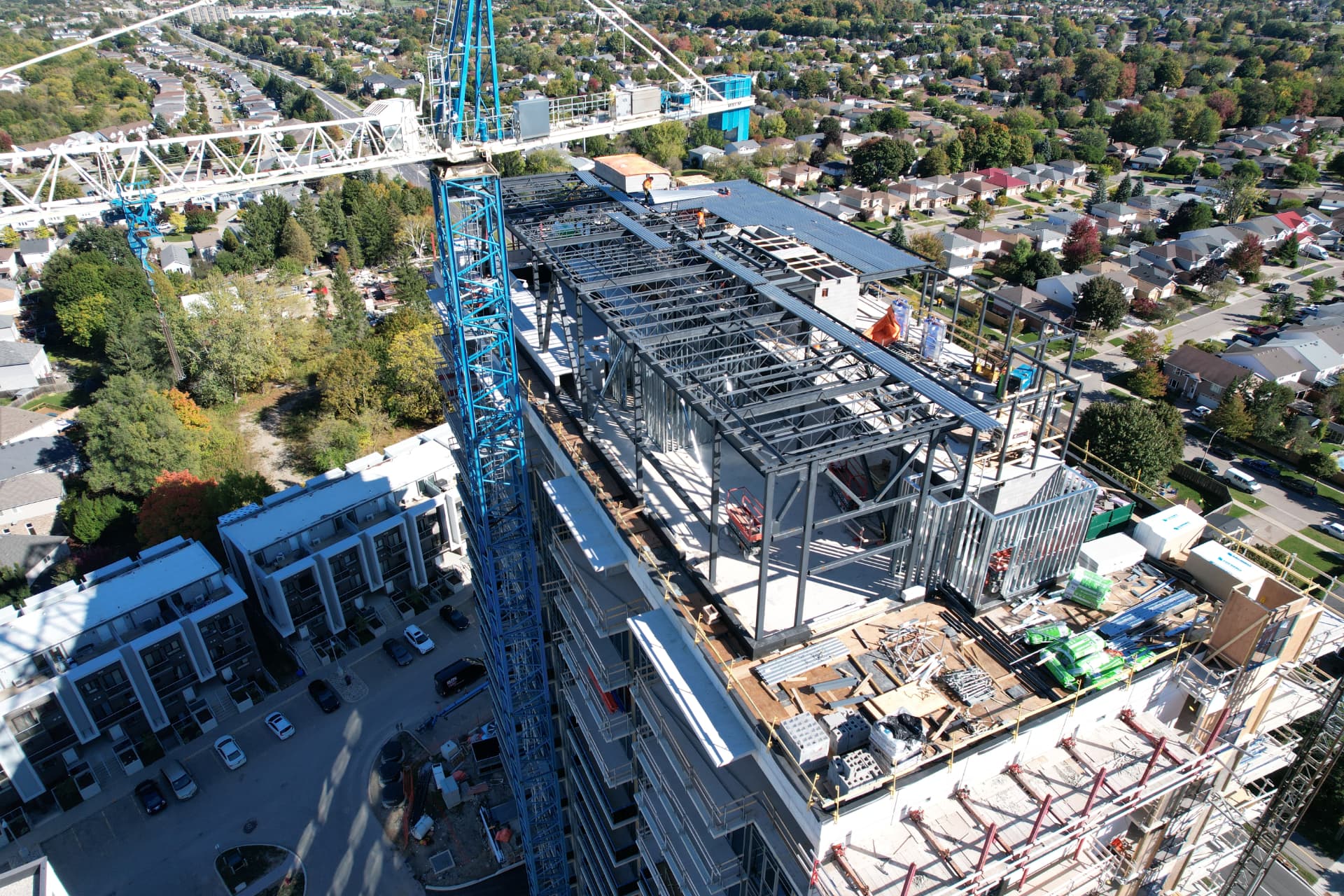 Rooftop penthouse steel framing with open-web joists and metal deck beside a tower crane — aerial view